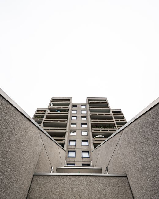 The image shows a view looking up between two grey concrete walls, which enclose a modern multi-storey residential building in the background. The building features several balconies with metal railings, some of which are partially decorated with plants or outdoor furniture. The structure has a symmetrical design with a central tower of uniformly sized windows, and the balconies are arranged in a tiered fashion on either side of this central core. The sky above is overcast, creating diffuse lighting that illuminates the scene evenly. The environment suggests urban residential surroundings, with the high-rise building indicating a densely populated area. The overall scene is neutral and muted in colour, with the concrete surfaces and metal railings contrasting lightly against the pale sky. This type of setting could imply the context of waste management needs associated with apartment complexes, where private or independent rubbish collection may be arranged, such as through services like Waste Disposal Ilford, to handle refuse removal efficiently from such multi-floor structures, especially when access for standard collection might be restricted or challenging.