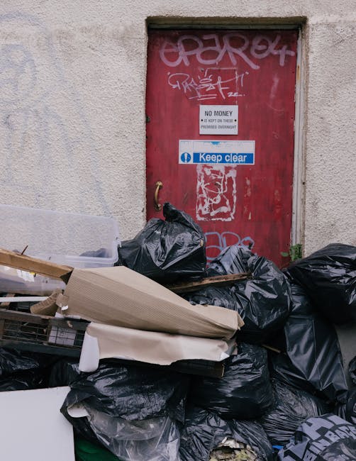 A pile of black garbage bags, some torn open with visible rubbish inside, located in front of a red metal door embedded in a concrete building wall. The door features various graffiti tags in white and pink spray paint, with a small white sign reading 'No Money' and a blue sticker with 'Keep clear' written on it. The rubbish bags and discarded cardboard boxes are positioned on a paved surface, near the corner of the building, suggesting an unmanaged waste collection site. The environment appears urban, with the scene indicating an area where private waste clearance or rubbish removal services, such as those provided by Waste Disposal Ilford, might be required due to the excess accumulation of refuse that obstructs access to the door, which could relate to access problems for flats situated above shops or similar properties. The photograph emphasizes the necessity for organized rubbish management and highlights the typical challenges of waste disposal in busy city areas, especially where on-site clearance is needed outside regular municipal services.