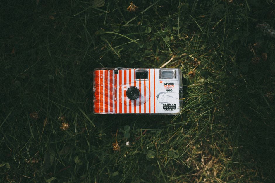 A compact, vintage-style disposable film camera with a primarily white body decorated with red and white vertical stripes, positioned on a patch of green grass with small plants and clover leaves. The camera features a black circular lens near the center, a small rectangular viewfinder at the top right corner, and a silver flash unit at the top left. The grass surrounding the camera appears natural and slightly uneven, with some brown and yellow blades interspersed among the green. The lighting suggests daylight, casting soft shadows and highlighting the camera's textured surface. The scene conveys a casual outdoor setting, where the camera may be part of a collection of miscellaneous items or waiting to be used for capturing moments, aligning with themes of personal photography and informal waste handling within a waste removal or disposal context that includes private and on-site clearance methods.
