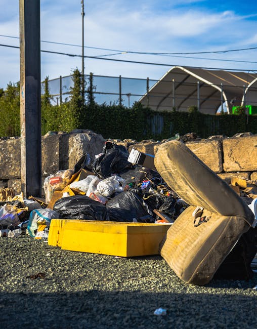 A pile of household waste and discarded items is scattered among dried, leafless branches on the ground in an outdoor setting. The debris includes various cardboard boxes, some partially collapsed or torn, with printed labels and brand markings visible. Among the cardboard, there are plastic containers, a blue plastic basket, small plastic toys, a black plastic drawer unit, and miscellaneous plastics with different textures and finishes. A wooden plank and a metal rod are also partially buried in the mixture of rubbish. The overall scene appears to be an unmanaged outdoor dump site, with waste items loosely piled in a natural environment with sparse vegetation in the background. The lighting is natural, suggesting daytime with overcast or diffuse light. The image illustrates the kind of waste that might be involved in private disposal or illegal dumping, which Waste Disposal Ilford can assist with through proper rubbish removal services to avoid fines for fly tipping and promote responsible waste management practices.