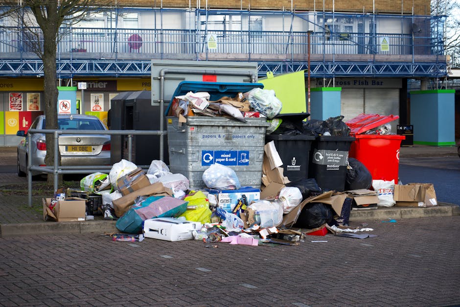 A large collection of mixed waste, including crumpled cardboard boxes, plastic bags, paper, and discarded packaging materials, is piled around multiple waste bins on a paved urban sidewalk. The bins comprise a grey bin labeled for mixed paper and card, a black bin for general waste, and a bright red bin possibly designated for other refuse or recyclables, with some lids open and rubbish overflowing. Surrounding the waste area, there are metal railings, a parked silver car with a partially visible yellow license plate, and a backdrop of a commercial building facade with storefronts, including a sign indicating a fish bar. In the background, a two-story structure features a blue metal scaffolding and safety barriers, hinting at ongoing maintenance or refurbishment work. The overall scene depicts an outdoor rubbish accumulation spot that might be linked to an independent waste collection service, such as waste disposal Ilford, emphasizing the importance of proper rubbish removal and disposal for urban environments.