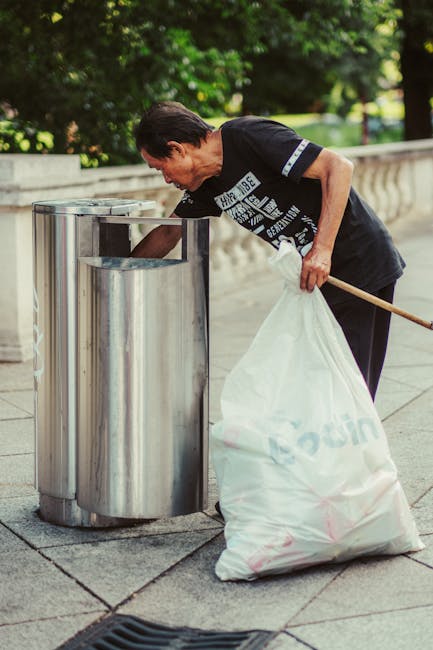 An elderly man wearing a black t-shirt with white text and dark trousers is seen using a metal public waste bin on a paved walkway. He is bent slightly forward, holding a large white rubbish bag with his left hand while inserting trash into the open-top, cylindrical, stainless steel bin with his right hand. The bin features a smooth, reflective surface and is positioned adjacent to another similar container, both set against a backdrop of lush green trees and a stone balustrade, indicating an outdoor park or urban environment. The scene is lit by natural daylight, highlighting the textures of the man's clothing, the metallic sheen of the bins, and the surrounding foliage. The cleanup activity reflects local or private waste disposal efforts often seen in community-maintained spaces, aligning with tasks undertaken by independent rubbish removal services such as Waste Disposal Ilford, based in Ilford. The overall composition emphasizes practical waste management activity within a peaceful outdoor setting, supporting the context of efficient rubbish collection and disposal handled outside standard municipal collection methods.