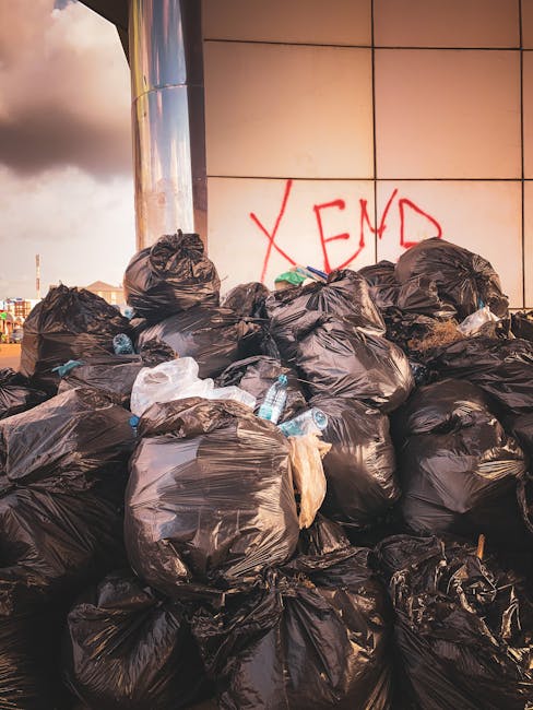 A large collection of mixed waste, including crumpled cardboard boxes, plastic bags, paper, and discarded packaging materials, is piled around multiple waste bins on a paved urban sidewalk. The bins comprise a grey bin labeled for mixed paper and card, a black bin for general waste, and a bright red bin possibly designated for other refuse or recyclables, with some lids open and rubbish overflowing. Surrounding the waste area, there are metal railings, a parked silver car with a partially visible yellow license plate, and a backdrop of a commercial building facade with storefronts, including a sign indicating a fish bar. In the background, a two-story structure features a blue metal scaffolding and safety barriers, hinting at ongoing maintenance or refurbishment work. The overall scene depicts an outdoor rubbish accumulation spot that might be linked to an independent waste collection service, such as waste disposal Ilford, emphasizing the importance of proper rubbish removal and disposal for urban environments.