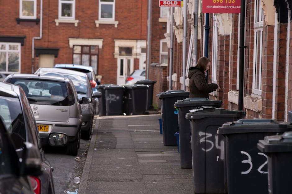 A residential street scene showing a row of dark grey wheelie bins placed on the pavement outside brick terraced houses, with some bins open and lidless. The bins are lined up along the edge of the sidewalk in front of the houses, which feature white window frames and decorative stone lintels. A person wearing a dark coat is standing next to one of the bins, appearing to be discarding rubbish. Several parked cars, including a silver hatchback and a darker vehicle, are positioned along the street facing away from the camera, with their windshields reflecting daylight. The background reveals additional brick houses and another row of bins further down the pavement. The environment is typical of a UK urban residential area, and the scene highlights the importance of proper waste disposal, possibly related to a private rubbish collection service such as Waste Disposal Ilford, in contrast to council-managed collection methods.