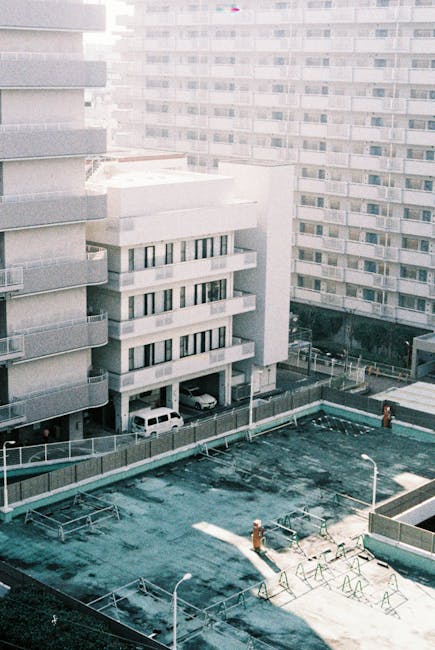 An outdoor scene showing a collection of overflowing rubbish bins and scattered waste on a paved area in front of a commercial building. The main visible bin is a large, grey mixed paper and card container with a partially open lid, overflowing with various paper, cardboard, and packaging materials. Surrounding this are several smaller black, green, and red bins, some of which are also overflowing with black refuse bags and loose rubbish, including plastic bottles, food wrappers, and cardboard boxes. The waste spills onto the surrounding pavement, with items such as a cardboard box, plastic bags, and paper parcels strewn on the ground. Behind the bins, there are storefronts with signage for local businesses and a building with a blue metal scaffolding structure or balcony. A parked silver car is visible to the left, partially obscured by a metal fence, and a tree with a bare trunk is on the near left side of the scene. The scene is captured in natural daylight with no visible people, indicating a typical urban rubbish accumulation possibly related to private waste collection or on-site clearance managed by waste disposal services like Waste Disposal Ilford.