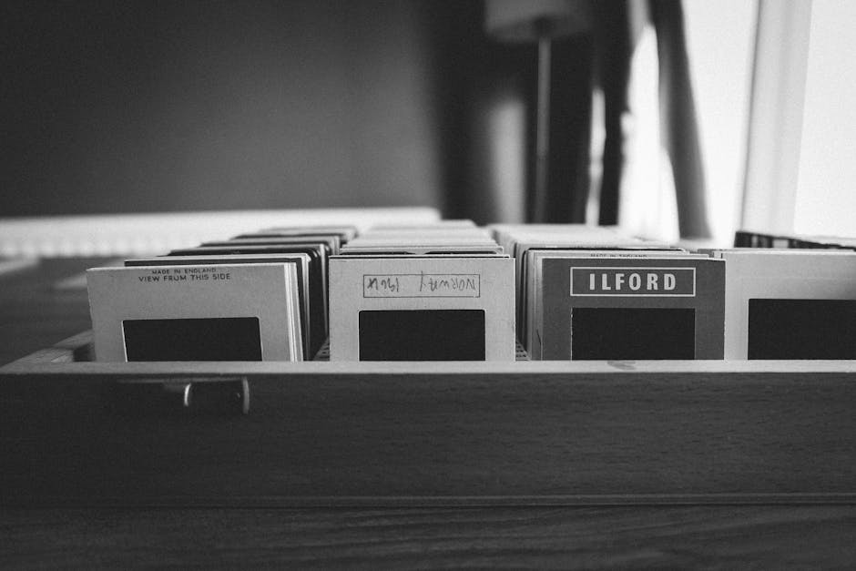 A black-and-white image showing a wooden tray or box containing neatly arranged rectangular flashcards or index cards, with some cards featuring bold labels such as 'ILFORD' and handwritten notes like 'NATIONAL UK'. The cards are upright, partially visible from the front, and appear to be made of smooth, thick paper or cardboard. The tray is positioned on a flat surface, likely a table or desk, with a background that includes blurred vertical elements and soft lighting, creating a neutral and professional atmosphere. The setting suggests an environment related to photo processing or documentation, subtly aligning with the theme of managing or organizing physical materials. Waste Disposal Ilford occasionally handles various waste types, including paper-based materials, in private or on-site clearance scenarios, making this scene relevant to waste management services provided.
