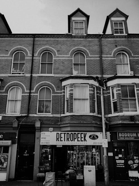 The image shows the facade of a three-story brick building with large arched windows on the second floor and smaller dormer windows on the third floor. The ground floor features a storefront with a sign reading 'RETROPEEK HOUSE CLEARANCES' in bold, black lettering on a white background, indicating a business specializing in private rubbish removal and house clearance services. Adjacent shop signs promote other local services, such as a beauty salon and a food delivery establishment, visible to the sides. The storefront includes glass doors and windows, with some equipment and items possibly related to waste or clearance activities situated outdoors in front of the shop. The building's exterior has a traditional brick finish with contrasting white trim around the windows and decorative brickwork. The scene appears to be in an urban setting, possibly on a busy street, with atmospheric overcast lighting contributing to a neutral tone. The overall environment suggests a commercial area where private waste disposal and rubbish removal services like those provided by Waste Disposal Ilford are available, supporting alternative rubbish collection methods outside of local authority services.