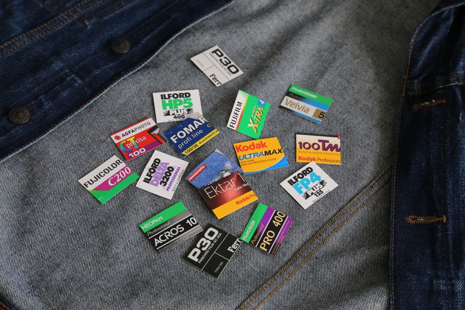 A close-up image of a section of denim fabric, likely from a pair of jeans, with various small photographic print labels scattered across the surface. The labels include different brand names, color indicators, and technical information such as 'Ilford HP5 Plus 400,' 'Kodak Ultra Max,' 'Fujicolor,' and 'Foma Profi Line,' among others. They feature a mix of bright colors, including green, yellow, red, purple, and black, with each label displaying distinct text and graphics, some with barcodes or numerical codes. The fabric background shows a textured, medium-wash blue denim with visible stitching along the right edge, and the labels rest on the fabric without any visible attachment or sticking. The scene is evenly lit, ensuring clarity of the labels' details and the fabric's texture, and the arrangement appears random, with some labels overlapping slightly. This image may be relevant in a context discussing photographic media, label identification, or a visual metaphor for sorting or categorising waste or materials, subtly aligning with rubbish removal or media disposal services offered by Waste Disposal Ilford.