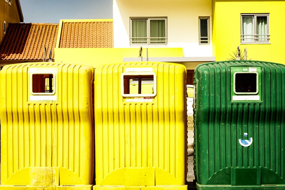 A row of three large, rectangular waste containers positioned outdoors on a paved surface in front of a modern, multi-storey residential building. The containers are made of ribbed plastic with a glossy finish, featuring small, rectangular openings in their upper sections for inserting rubbish. Two of the containers are bright yellow, with one showing some visible dirt marks and scuffing, while the third container is dark green with a slightly worn appearance and a small, circular symbol on its lower front. The building behind them has a white and neon yellow facade, with windows and balconies visible, and the roof is covered with reddish-brown tiles. The scene is illuminated by natural daylight, highlighting the vibrant colors of the containers and building, illustrating a typical setting for private waste collection or rubbish removal services managed by Waste Disposal Ilford in a suburban area.