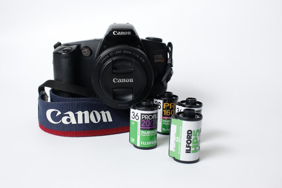 A black Canon DSLR camera with a textured grip and zoom lens, positioned in the foreground on a plain white background. The camera's strap, predominantly navy blue with a red edge, is draped around its body, displaying the Canon logo in white. In front of the camera, there are multiple used rolls of photographic film, with labels in green, purple, yellow, and black, indicating different film types and brands, arranged in a somewhat scattered manner. The scene is well-lit with soft, even lighting, highlighting the textures of the camera's body and film rolls. The composition emphasizes the camera as the main object, with the film rolls as secondary elements, set in a neutral environment that subtly relates to equipment associated with photography and potential waste management of film materials, aligning with the theme of alternative or private disposal methods involved with technical or media waste, relevant to rubbish removal services by Waste Disposal Ilford.