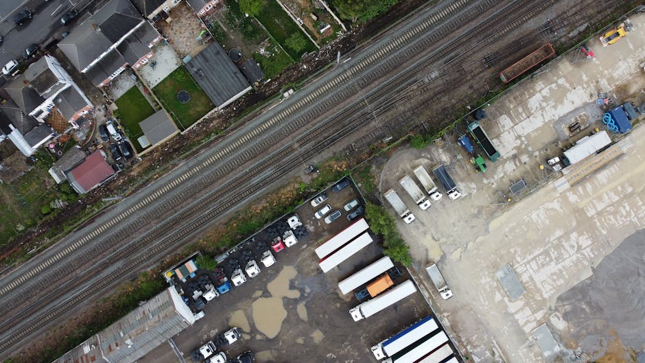 An aerial view of a mixed-use area showing a railway track running horizontally across the upper part of the image, with gravel and ballast visible beneath the rails. Below the railway, there is a paved lot filled with various vehicles including cars, vans, and trucks, some of which appear to be used for commercial or waste management purposes. The vehicles are parked in an irregular pattern, with some adjacent to stackable metal storage containers and others near a cluster of stacked white and blue cargo trailers. To the left, there are residential houses with brick exteriors, small gardens, and rooftops in shades of gray and red, partially bordered by fences and greenery. The scene is lit by natural daylight, with some areas showing damp patches or puddles, possibly from recent rain, emphasizing the utilitarian nature of the space. The setting reflects an environment used for private logistics, storage, or waste disposal activities, relevant to rubbish removal and alternative waste handling services, with the presence of company-related vehicles and equipment relevant to waste management companies like Waste Disposal Ilford.