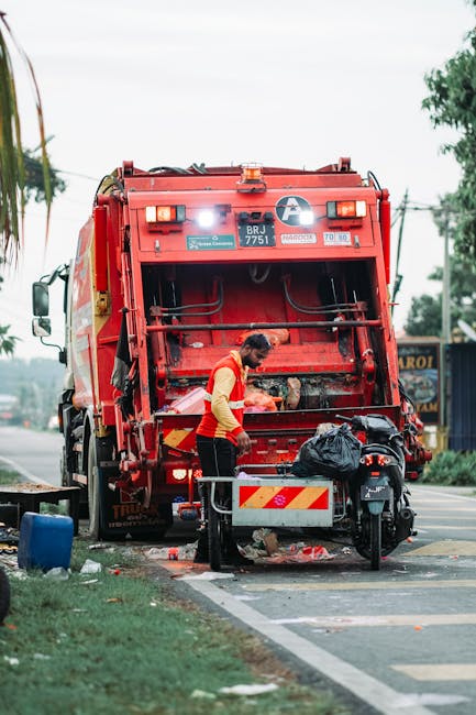 A waste collection operative from Waste Disposal Ilford is seen working at the rear of a large, red rubbish truck on a roadside. The truck's rear compactor panel is open, revealing mechanical and hydraulic components made of metal with a slightly worn, industrial finish. The worker, dressed in a yellow and red high-visibility vest and dark trousers, is leaning forward, handling black plastic rubbish bags that are placed in a designated area at the back of the vehicle. The scene is set on a paved street with a white line marking the edge of the parking or loading zone. To the right of the worker, there is a motorcycle parked adjacent to a small, waist-high metal barrier with red and white reflective stripes. In the immediate background, there are utility poles, wires, and blurred trees, suggesting a suburban or urban environment. The lighting appears natural and overcast, consistent with daytime. This image exemplifies an on-site waste removal process, which may be part of private waste handling services provided by Waste Disposal Ilford as an alternative to council rubbish collection, emphasizing efficient and professional local rubbish management.